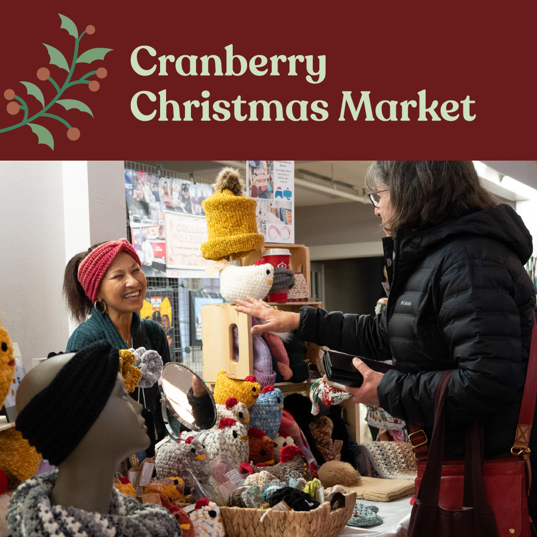 A vendor smiles warmly at a customer at a holiday market stall filled with handmade crochet items and crafts. The event title, "Cranberry Christmas Market," is displayed in white text on a deep red background at the top.