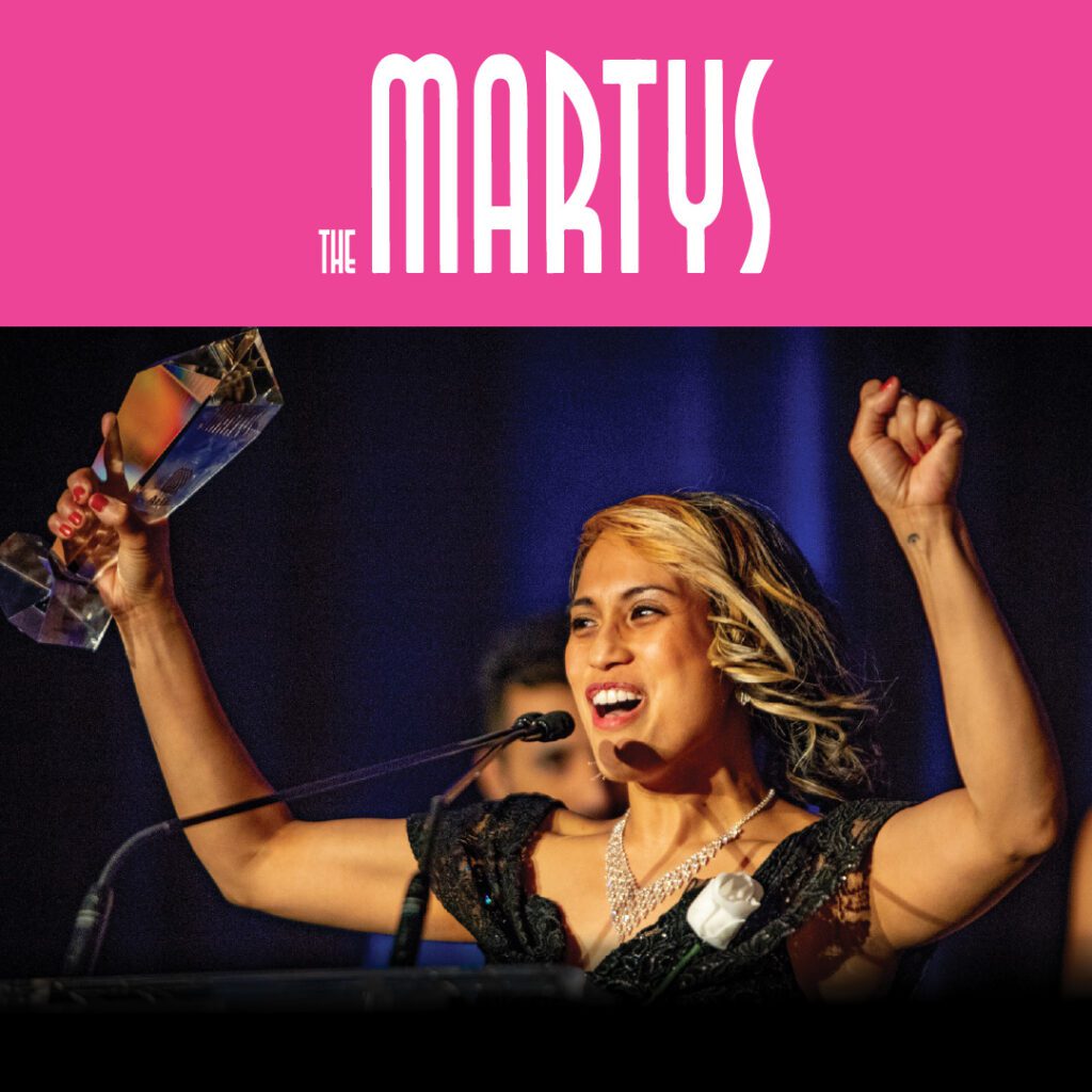 A woman on stage, holding a MARTY award trophy high in one hand and raising the other in celebration. The event title, "The MARTYS," is displayed in pink text at the top.