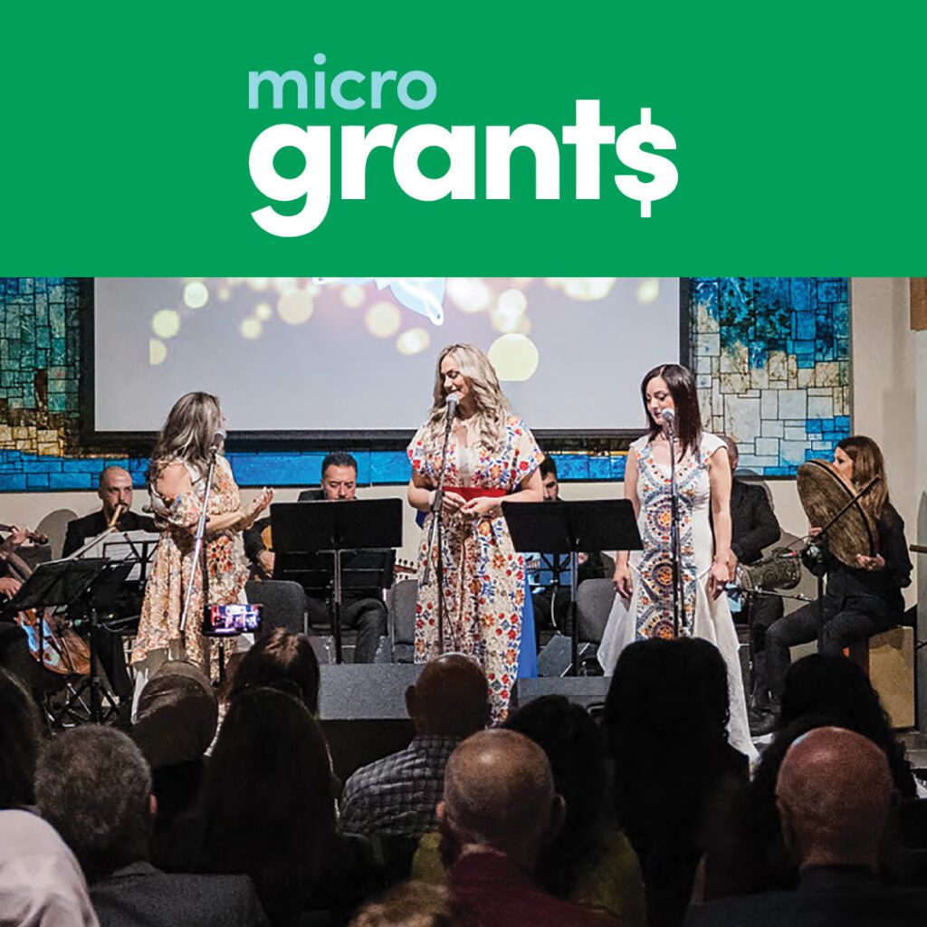 A live performance featuring three women in traditional-style dresses singing on stage with musicians playing instruments behind them. The audience watches attentively in an intimate venue with warm lighting