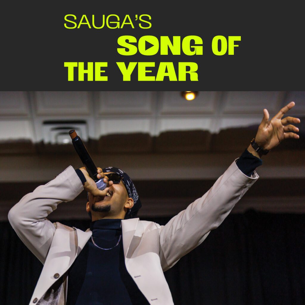 A performer on stage sings into a microphone, dressed in a stylish beige jacket and a bandana, raising one hand in the air. The event title, "Sauga’s Song of the Year," is displayed in neon green text at the top.